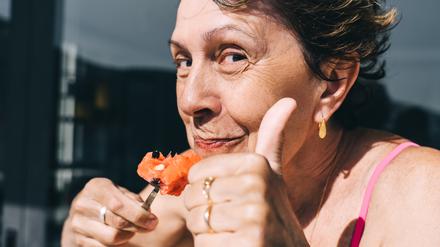 Eine ältere Dame isst eine Wassermelone und zeigt einen Daumen nach oben.