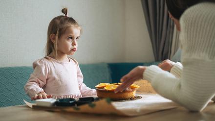 Eine Mutter präsentiert ihrer etwa sechsjährigen Tochter am Esstisch erwartungsvoll einen Kuchen.
