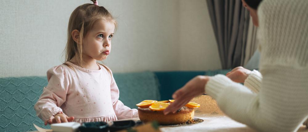Eine Mutter präsentiert ihrer etwa sechsjährigen Tochter am Esstisch erwartungsvoll einen Kuchen.
