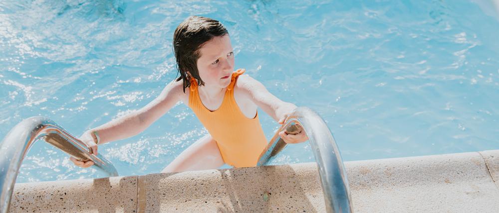 Ein Mädchen steigt aus einem Pool und blickt dabei verunsichert über die Schulter. Es trägt einen orangefarbenen Badeanzug.
