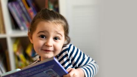 Ein etwa vierjähriges Mädchen hält ein Buch in der Hand und schaut den Betrachter mit verschmitztem Lächeln an. 