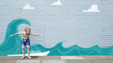 Ein etwa sechsjähriger Junge in Badehose steht in Surferhaltung vor einer Wand, die mit Wellen bemalt ist.