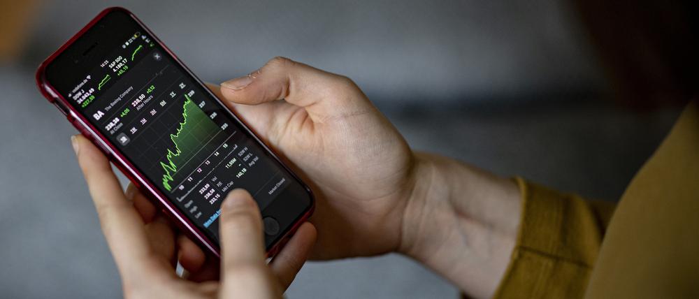 Hände halten ein Smartphone, auf dem ein Aktienchart abgebildet ist.