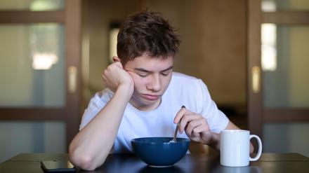 Ein Teenager sitzt mit geschlossenen Augen an einem Tisch und hält dabei einen Löffel in eine Müslischüsssel.
