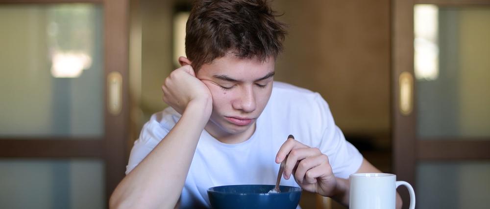 Ein Teenager sitzt mit geschlossenen Augen an einem Tisch und hält dabei einen Löffel in eine Müslischüsssel.
