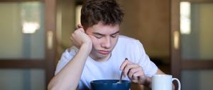 Ein Teenager sitzt mit geschlossenen Augen an einem Tisch und hält dabei einen Löffel in eine Müslischüsssel.
