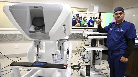 Der US-amerikanische Chirurg Vipul Patel steht neben einem OP-Roboter. Im Hintergrund ist auf einem Bildschirm das per Telemedizin zugeschaltete angolanische OP-Team zu sehen. 