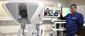 Der US-amerikanische Chirurg Vipul Patel steht neben einem OP-Roboter. Im Hintergrund ist auf einem Bildschirm das per Telemedizin zugeschaltete angolanische OP-Team zu sehen. 