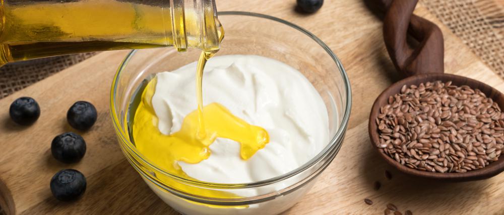 Flax seed oil is being poured over cottage cheese in a bowl, with flax seeds and blueberries in the background
