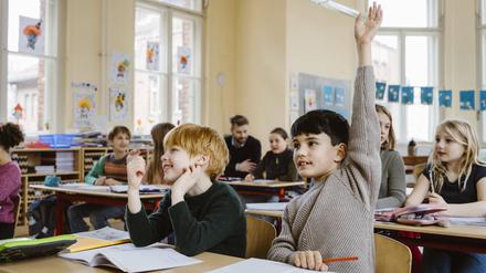 Zwei Jungen sitzen an einem Tisch im vollen Klassenzimmer. Einer meldet sich mit hochgerecktem Arm, der andere nicht.