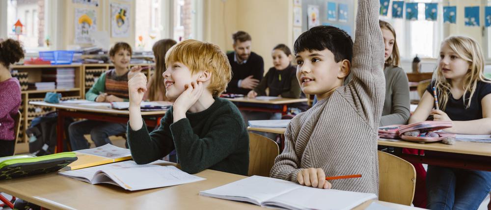 Zwei Jungen sitzen an einem Tisch im vollen Klassenzimmer. Einer meldet sich mit hochgerecktem Arm, der andere nicht.