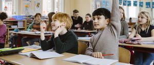 Zwei Jungen sitzen an einem Tisch im vollen Klassenzimmer. Einer meldet sich mit hochgerecktem Arm, der andere nicht.