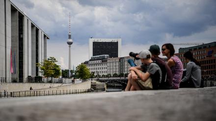 Touristen sitzen am Spreeufer in der Nähe des Bundestags. Im Hintergrund ragt der Fernsehturm am Alexanderplatz in die Höhe. +++ dpa-Bildfunk +++