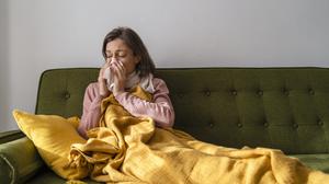 Eine Frau sitzt mit Decke auf dem Sofa und schnäuzt sich die Nase.