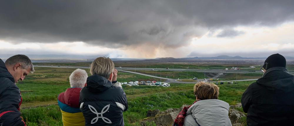 FILES) Touristen, die am 16. Juli 2025 neben einem Denkmal in Keflavik, Island, stehen, beobachten aus der Ferne Rauch und Lava, die nach dem Ausbruch des Vulkans in der Nähe von Grindavik auf der isländischen Halbinsel Reykjanes ausströmen.