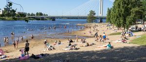 Menschen genießen einen heißen Tag am Fluss Kemijoki, neben der Lumberjack’s Candle Bridge, in Rovaniemi, Finnland.