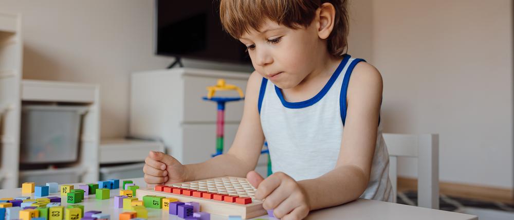 Ein etwa drei bis vier Jahre altes Kind mit kurzen Haaren sitzt an einem Tisch und beschäftigt sich konzentriert mit bunten Spielsteinen, auf den Zahlen abgebildet sind.