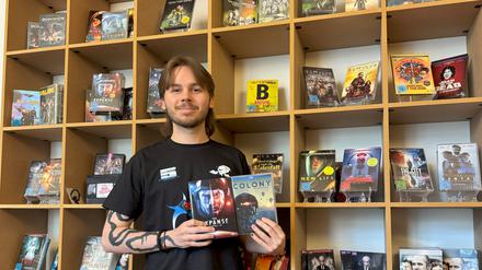 Der Auszubildende Lukas steht vor einem Regal mit DVDs und hält eine davon in die Kamera.