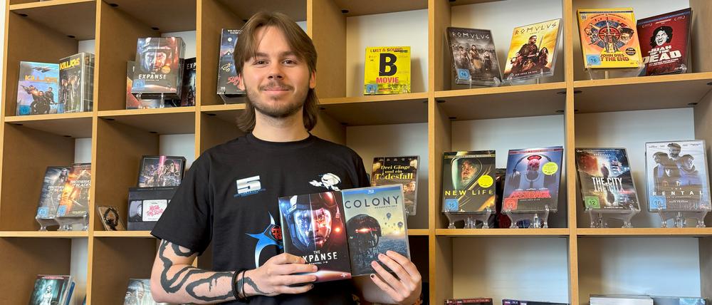 Der Auszubildende Lukas steht vor einem Regal mit DVDs und hält eine davon in die Kamera.