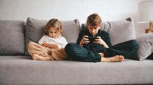 Zwei Kinder sitzen auf einer grauen Couch und starren jeweils auf ein Smartphone.