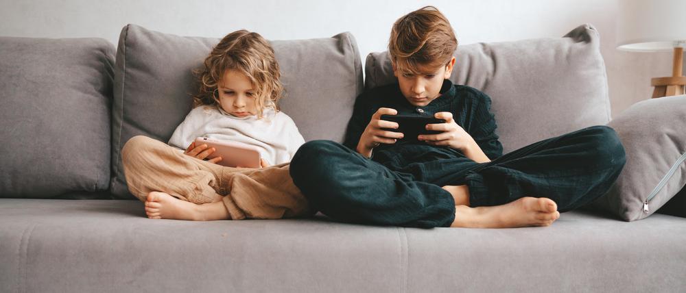 Zwei Kinder sitzen auf einer grauen Couch und starren jeweils auf ein Smartphone.