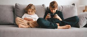 Zwei Kinder sitzen auf einer grauen Couch und starren jeweils auf ein Smartphone.