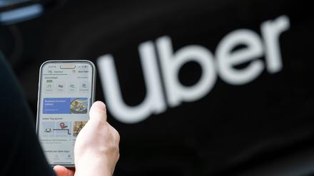 Ein Mann steht vor einem Mietwagen mit einem Schriftzug des Mobilitätsdienstleisters und Fahrtenvermittlers Uber und hält sein Smartphone mit der App des Unternehmens in der Hand (Symbolbild).