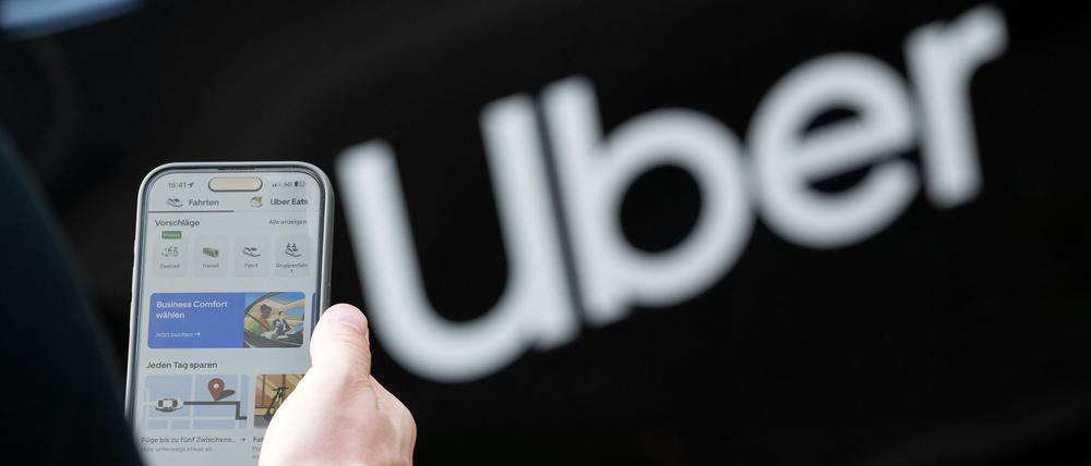 Ein Mann steht vor einem Mietwagen mit einem Schriftzug des Mobilitätsdienstleisters und Fahrtenvermittlers Uber und hält sein Smartphone mit der App des Unternehmens in der Hand (Symbolbild).