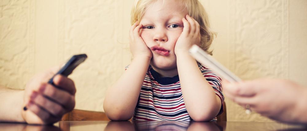 Eltern tippen, wischen, lesen, während das Kind aufblickt und wartet. Was alltäglich wirkt, kann die frühe Entwicklung von Kindern stören.