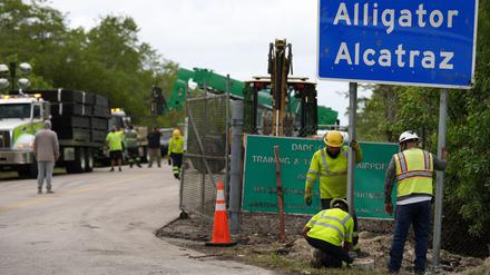 Arbeiter installieren ein Schild mit der Aufschrift «Alligator Alcatraz» am Eingang einer neuen Haftanstalt für Migranten an der Dade-Collier-Ausbildungs- und Übergangseinrichtung, während in der Nähe große Zaunplatten von einem Laster abgeladen werden. 