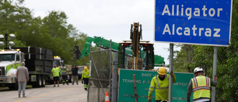 Arbeiter installieren ein Schild mit der Aufschrift «Alligator Alcatraz» am Eingang einer neuen Haftanstalt für Migranten an der Dade-Collier-Ausbildungs- und Übergangseinrichtung, während in der Nähe große Zaunplatten von einem Laster abgeladen werden.