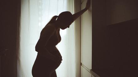 Eine schwangere Frau, die eher als Silhouette vor einem Fenster zu sehen ist, stützt sich mit der Hand an einem Schrank ab, um eine Wehe besser zu ertragen  (Symbolbild).
