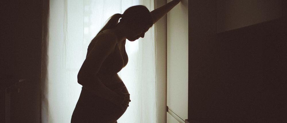 Eine schwangere Frau, die eher als Silhouette vor einem Fenster zu sehen ist, stützt sich mit der Hand an einem Schrank ab, um eine Wehe besser zu ertragen  (Symbolbild).