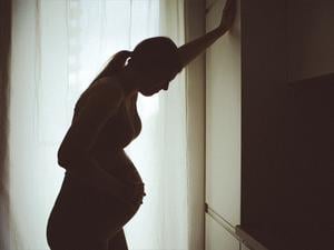 Eine schwangere Frau, die eher als Silhouette vor einem Fenster zu sehen ist, stützt sich mit der Hand an einem Schrank ab, um eine Wehe besser zu ertragen  (Symbolbild).