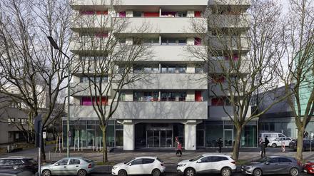 Ein massives, sechsstöckiges Wohngebäude mit roten Sonnenläden und Balkonen sowie zwei großen Eingangspfeilern steht an einer Straße, an der Autos parken. Im Erdgeschoss ist die Fassade verglast.