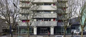 Ein massives, sechsstöckiges Wohngebäude mit roten Sonnenläden und Balkonen sowie zwei großen Eingangspfeilern steht an einer Straße, an der Autos parken. Im Erdgeschoss ist die Fassade verglast.