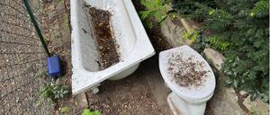 Eine von Laub und Regenwasser verschmutzte Toilette und Badewanne stehen auf einer Terrasse zwischen Bäumen und einem Gartenzaun. 