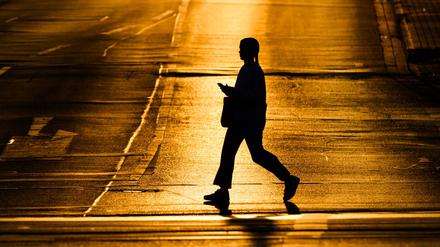 Eine Frau läuft mit einem Smartphone in der Hand über eine Straße, die von der aufgehenden Sonne in warmes Licht getaucht wird.