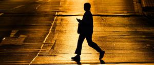 Eine Frau läuft mit einem Smartphone in der Hand über eine Straße, die von der aufgehenden Sonne in warmes Licht getaucht wird.