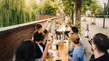 Menschen sitzen an einem langen Biertisch auf dem Gehweg und stoßen an. 