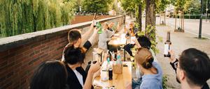 Menschen sitzen an einem langen Biertisch auf dem Gehweg und stoßen an. 