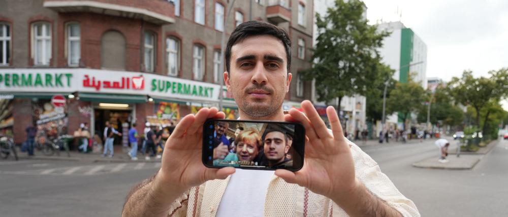 Anas Modamani steht auf der Sonnenallee in Berlin. Der Syrer kam 2015 als Flüchtling nach Deutschland. Er zeigt auf seinem Handy ein Selfie mit der damaligen Bundeskanzlerin Angela Merkel.