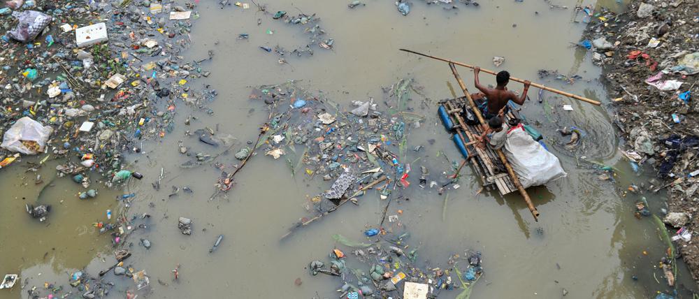Bangladesch: Ein Vater rudert mit seinem Sohn auf einem handgefertigten Floß und sucht im mit Plastikmüll gefüllten Kanal von Kazir Bazar in Sylhet nach Recyclingprodukten aus Plastik.