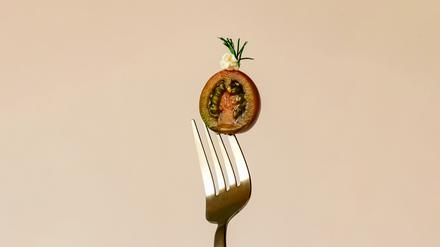 Eine kunstvoll drapierte, halbierte Tomate ist von einer Gabel aufgespießt vor pastellig-orangen Hintergrund zu sehen. (Symbolbild)
