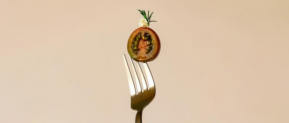Eine kunstvoll drapierte, halbierte Tomate ist von einer Gabel aufgespießt vor pastellig-orangen Hintergrund zu sehen. (Symbolbild)