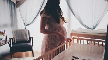 Eine schwangere Frau steht allein an einem Babybett, in dem zwei Kuscheltiere liegen.