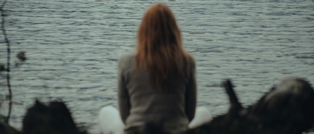 Eine Frau mit langen Haaren blickt auf ein Gewässer. Sie ist nur von hinten zusehen.