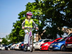 Eine Grundschülerin fährt auf ihrem Fahrrad bei einer Grundschule vor parkenden Autos entlang.