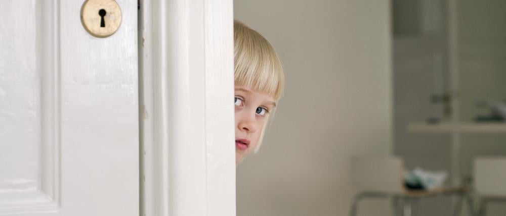 Ein scheues Kind versteckt sich hinter einer Tür.