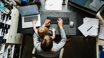 Ein erwachsener Mensch mit kurzen Haaren sitz an einem Schreibtisch und arbeitet am Computer. Auf dem Schoß hat er oder sie ein jüngeres Kind. Das Foto ist von oben aufgenommen, sodass man die Gesichter nicht sehen kann.